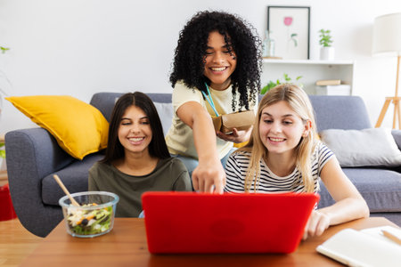 Cheerful young female students eating takeaway food and using laptop at homeの写真素材
