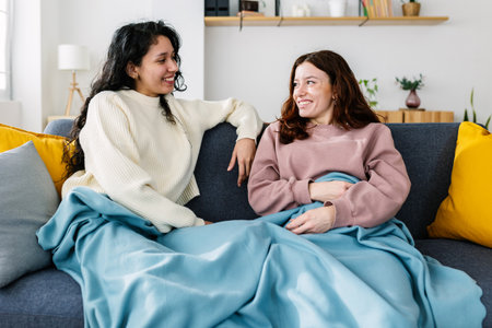 Two young smiling latin american women covered with a blanket talking on sofaの写真素材