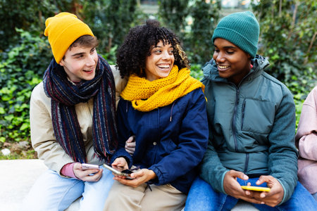 Diverse young students using mobile phone sitting on city bench on winter