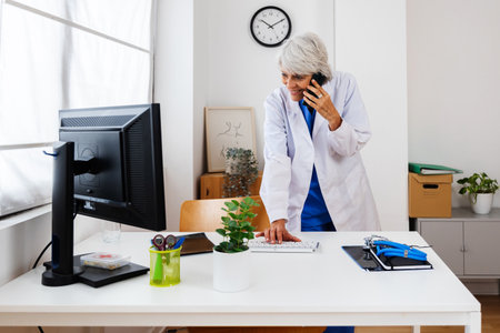 Senior female doctor talking on phone standing at deskの写真素材