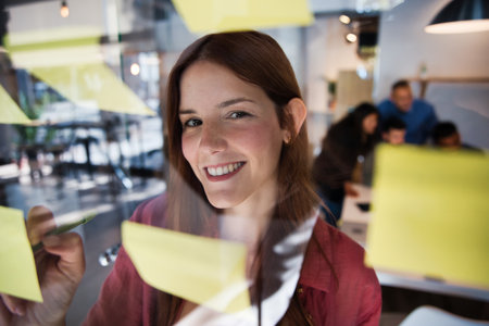 Smiling young woman writing notes on glass board during creative meetingの写真素材