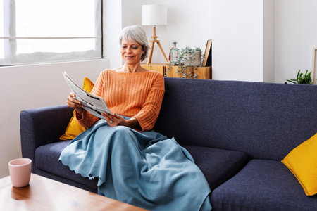 Smiling senior woman reading newspaper on sofa at homeの写真素材