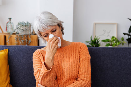 Senior woman with a cold blowing her nose on the couch at homeの写真素材
