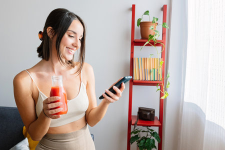 Young healthy woman using mobile phone while drinking tomato juice at homeの写真素材