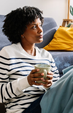 Young black woman relaxing on sofa drinking warm hot drink at home