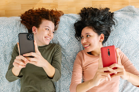 Top view of two young women having fun mobile phones lying together on bed.の写真素材