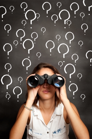 young woman using binoculars in front of chalkboard with interrogation symbolsの写真素材