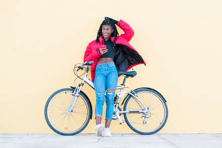 happy african-american woman wearing red jacket with bike on street over yellow background.の写真素材