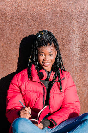 Beautiful African woman wearing red jacket with headphones outdoors. Black woman writing in a...