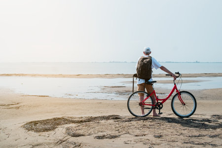 rear view of man with backpack standing and holding bike on seafrontの写真素材