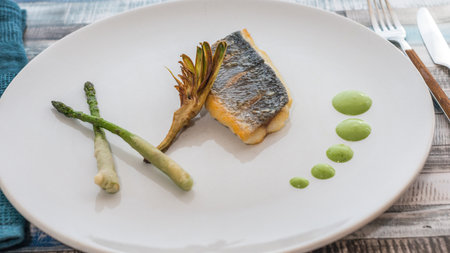 Steam bream sided by wild asparagus, artichoke and by dots of Dutch green sauce on a colored wooden table with cutleryの写真素材