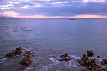 Sailboat on French riviera coast on moody sunset view. Town of Antibes waterfront. France.の写真素材