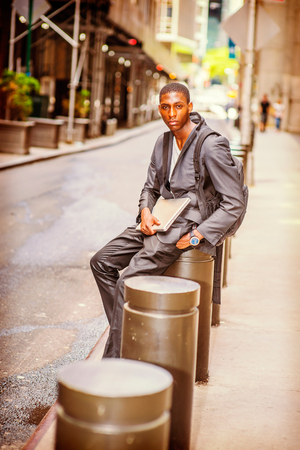 Young African American College Student carrying shoulder bag, holding laptop computer, sitting on pillar on vintage street in New York, thinking, lost in thought.の写真素材