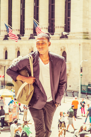 Young African American Man traveling in New York,  carrying shoulder bag, walking up on steps, standing in front of vintage office building, looking away.の写真素材