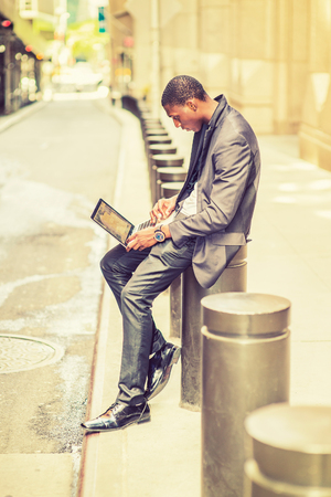 Working anywhere, anytime. Young man working on street. A young black college student is sitting outside an office building, typing on a laptop computer, reading, thinking. Retro filtered look.の写真素材
