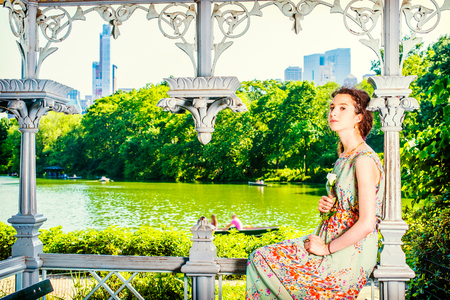 Young Lady Waiting for You. Dressing in sleeveless, long dress, a pretty teenage girl is sitting inside a pavilion, hands holding a white rose, looking at you. people rolling boats in background,.の写真素材