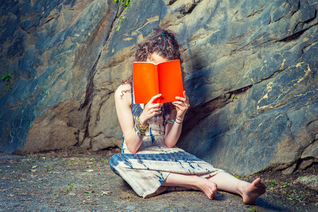 Girl Thinking Hard. Wearing long dress, bracelet, barefoot, a pretty teenage college student with curly long hair is sitting on ground against rocks, holding a red book covering her face, thinking.の写真素材