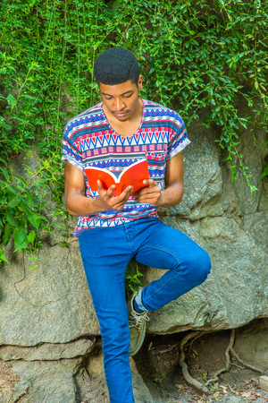 Man Reading Outside. Wearing a short sleeve, collarless, colorful pattern shirt, blue jeans, a young college student is sitting against rocks with green leaves, looking down, reading a red book,の写真素材