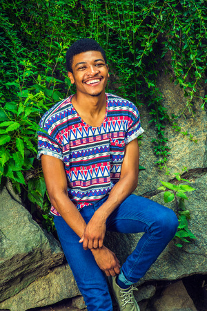 Portrait of Young Black Man. Wearing a short sleeve, collarless, colorful pattern shirt, a young handsome guy is sitting against rocks with green leaves, smiling, charmingly looking at you.の写真素材