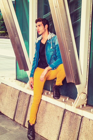 Man Missing You. Dressing in a blue jacket with hood, black under wear, yellow pants, leather boot shoes, a young handsome guy is standing by a metal structure on the corner, looking up, thinking.の写真素材