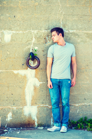 Man Missing You. Wearing gray T shirt, blue jeans, white sneakers, a young handsome guy is standing against an old concrete wall with a rusty metal ring, looking down at white rose, lost in thought.の写真素材