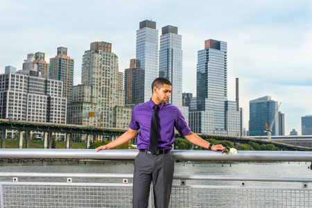 Dressing in a purple shirt, gray pants and a black tie, holding a white rose, a young guy with a little beard and mustache is thinking about you. The background is busy high buildings.の写真素材