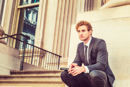 Man Waiting for You. Dressing in black suit with Shawl Lapel, black necktie, a young sexy guy with curly hair is squatting outside office, holding a laptop computer, relaxing.の写真素材