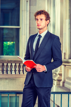 Man Reading Outside. Dressing in black suit with Shawl Lapel, black necktie, a young sexy guy with curly hair is standing inside office building, hands holding a red book, reading, looking forward.の写真素材