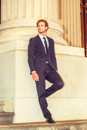 Man Waiting for You. Dressing in black suit with Shawl Lapel, black necktie, a young sexy guy with curly hair is standing by column outside office, bending a leg, looking away.の写真素材