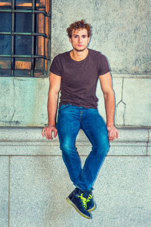 Man Relaxing Outside. Wearing dark brown T shirt, blue jeans, black sneakers, a young sexy guy with curly hair is sitting against wall by window, crossing feet, thinking, lost in thought.の写真素材