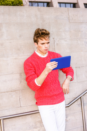 Young blonde professional, wearing red knit sweater, reading small tablet computer, standing on street against wall outside in windy day in New York. Reading anywhere.の写真素材