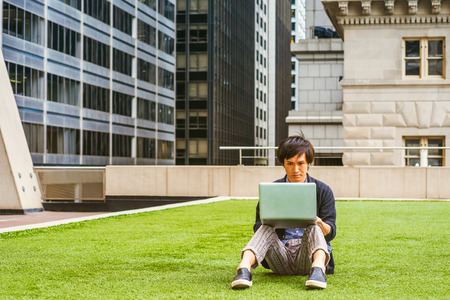 Wearing patterned shirt, black sweater, casual pants, leather shoes, a young guy sitting on green lawn in business district, reading, working on laptop computer.の写真素材