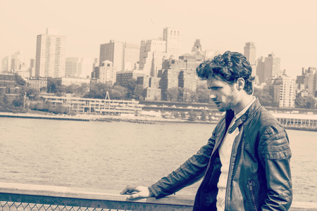 European businessman traveling on East River in New York. Young guy with beard, looking down, frowned, sad, tired, thinking, missing family, friends, home. Crowed building in Brooklyn on background.の写真素材