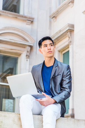 Young Asian American Man studying, working in New York, wearing gray blazer, white pants, sitting...