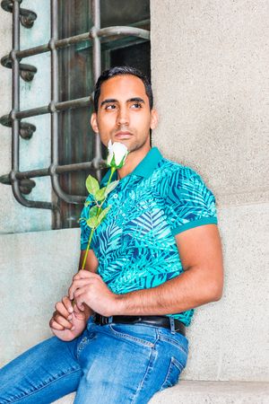 Young Hispanic American Man missing you, waiting for you, wearing green patterned Polo shirt, holding white rose, sitting against old wall with window on street in New York, looking away, thinking.の写真素材