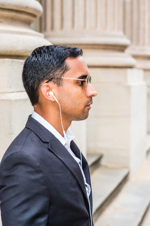 Young Handsome Hispanic American Businessman thinking outside in New York, wearing sunglasses, white wired earphone, standing by column of old office doorway, seriously listening music. Side View.の写真素材