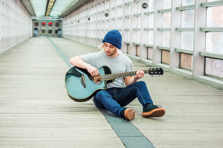 Young college student playing music, with long blonde hair, wearing knitted hat, gray T shirt, jeans, sneakers, sitting on wooden floor inside indoor walkway on campus in New York City, playing guitarの写真素材