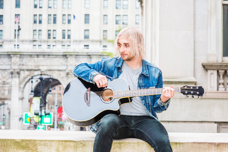 Musician Playing Guitar outdoor in New York City. Young handsome man with long blonde hair, wearing blue Denim jacket, black pants, gray shirt, sitting on street, playing guitar, looking dow, think.の写真素材