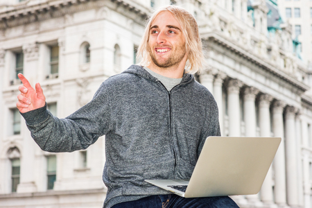 Man Working Outside. Young hipster college student with long blonde hair, wearing gray sweater with hood, sitting outside old style office building in New York, working on laptop computer, waving handの写真素材