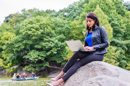 Young American Woman traveling, working in New York, wearing black leather jacket, black trousers, sitting on rocks by lake at Central Park, typing on laptop computer. People rowing boat on background.の写真素材