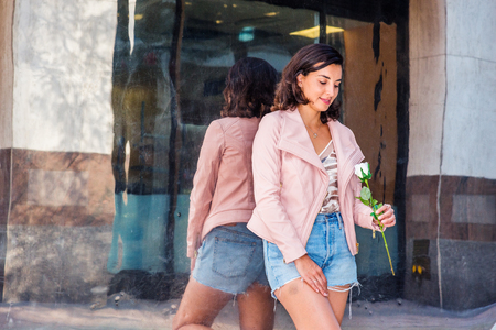 Young American Woman wearing light pink leather jacket, blue ripped Denim shorts, standing by metal mirror on street in New York, lowering head, looking at white flower held by hand, smiling, thinkingの写真素材