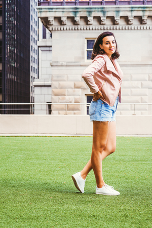 Portrait of Young American Female College Student in New York City, wearing light pink leather jacket, blue ripped Denim shorts, white sneakers, hands on hips, standing on green lawn on campus.の写真素材