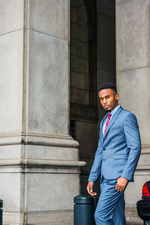 Young African American Businessman with beard traveling, working in New York, wearing dark sky blue suit, white striped shirt, violet red patterned tie, walking by columns on vintage street, looking.の写真素材