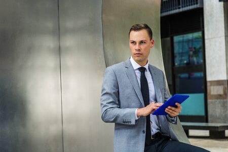 Modern Reading. Young Businessman wearing gray blazer, white shirt, black tie, standing by silver...