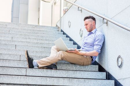 Way to Success. Young American college student studying in New York City, wearing light purple shirt, beige pants, black shoes, sitting on stairs, looking down, reading, working on laptop computer.の写真素材