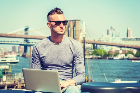 Young handsome American man traveling, working in New York City, wearing gray long sleeve T shirt, sunglasses, sitting by river, working on laptop computer. Brooklyn, Manhattan bridges on background.の写真素材