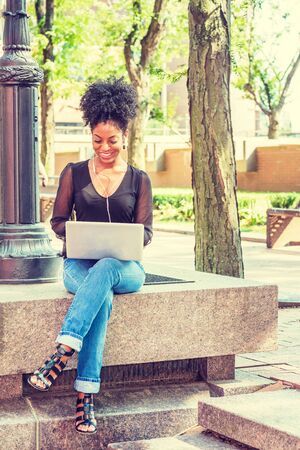 Young African American Woman with afro hairstyle wearing mesh sheer long sleeve shirt, jeans, sitting by light pole on campus in New York, listening music with earphone, working on laptop computer.の写真素材