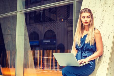 Young Eastern European American Woman working in New York, wearing blue sleeveless jumpsuit, standing against column outside office building, bending leg, working on laptop computer, looking away.の写真素材