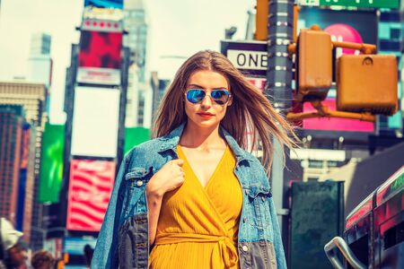 Young Eastern European Woman traveling in New York, with long brown hair, wearing yellow dress, blue Denim jacket draped over shoulder, blue sunglasses, standing on street in Times Square of Manhattan.の写真素材