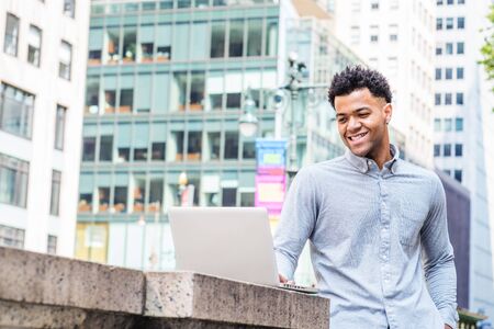 Young Mix-Race American Man working outside in New York City, wearing light gray long sleeve shirt, standing by half wall on street in Manhattan, reading on laptop computer, looking down, smiling.の写真素材